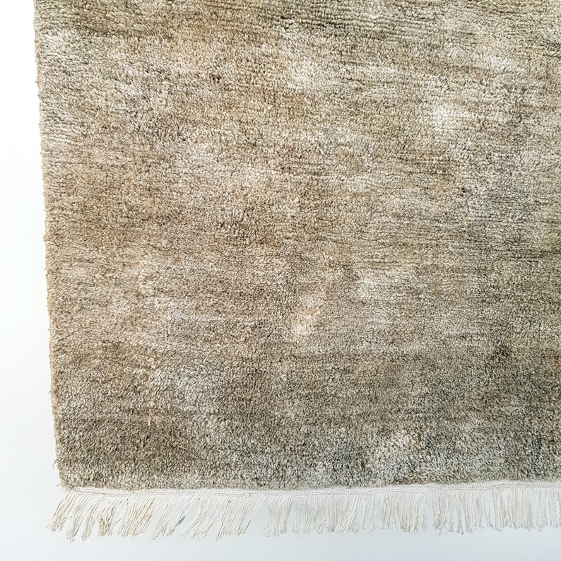 Op de afbeelding: Close-up van een beige tapijt met een getextureerd oppervlak. Het tapijt heeft een franje van witte kwastjes langs &eacute;&eacute;n rand. De kleur van het tapijt varieert van licht- tot donkerbeige.