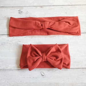 May include: Two rust-colored fabric headbands. One headband is tied in a simple knot, while the other is tied in a large bow.