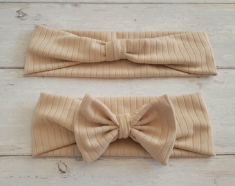 May include: Two beige ribbed fabric headbands with a bow. The headband on the top has a simple knot, while the headband on the bottom has a large bow.