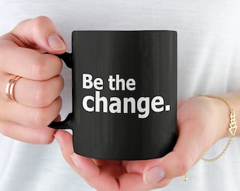 May include: Black ceramic mug with the text "Be the change" in white. The mug is held by a person wearing a white shirt. A gold bracelet and rings are also visible.