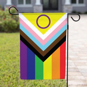 May include: A garden flag with a rainbow pride flag design, featuring a black and brown chevron pattern at the top, a yellow circle with a purple ring in the center, and a rainbow stripe pattern at the bottom.