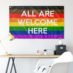 Puede incluir: Una bandera arco&iacute;ris del orgullo con el texto "ALL ARE WELCOME HERE" en letras blancas.