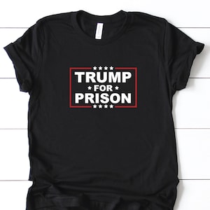 May include: Black t-shirt with the text "TRUMP FOR PRISON" in white, inside a red rectangular border, with white stars above and below the text. The shirt is displayed on a white wooden surface.