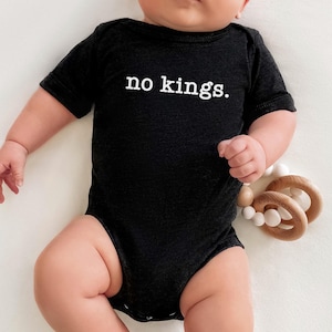 May include: A black baby bodysuit with the text "no kings." The short-sleeved onesie is made of a soft material. A wooden teething toy with white beads and wooden rings is next to the baby.