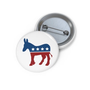 May include: A white button with a blue and red donkey silhouette. The donkey has five white stars on its back.