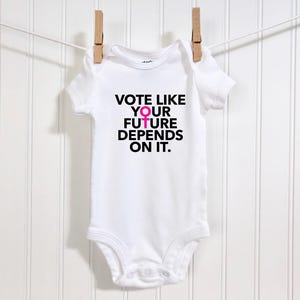 Puede incluir: Body de bebé blanco con el texto "VOTE LIKE YOUR FUTURE DEPENDS ON IT." en negro y rosa. El body está colgado de un tendedero con pinzas de madera sobre un fondo blanco.