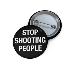 May include: A black button with the text "STOP SHOOTING PEOPLE" in white. The button is round and has a metal pin on the back. Another button is partially visible in the background.