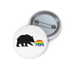 May include: A white button with a black silhouette of a bear and a rainbow bear silhouette. The rainbow bear is smaller and is facing the black bear.