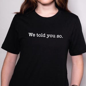 Puede incluir: Camiseta negra de cuello redondo con el texto blanco "We told you so." La camiseta de manga corta está hecha de un material suave. La camiseta es una prenda informal.