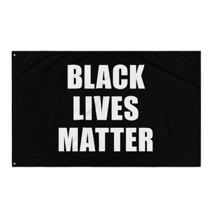 May include: Black flag with the words "BLACK LIVES MATTER" in bold white capital letters. The flag is rectangular with grommets in the corners for hanging. The background is solid black.