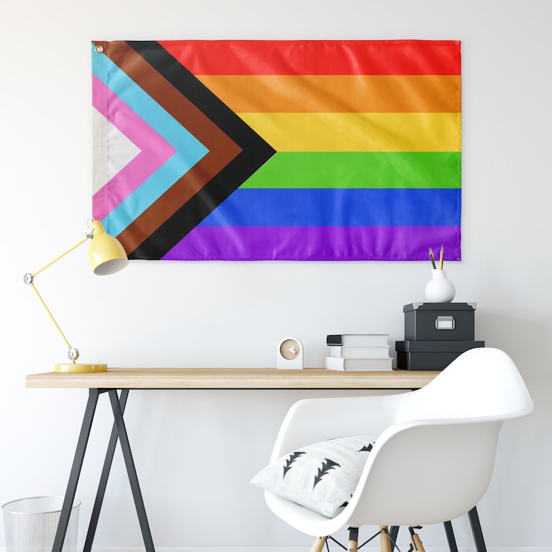 May include: A pride flag with a black, brown, light blue, pink, white, green, blue, and purple stripe pattern. The flag is hanging on a white wall above a desk with a lamp, a chair, and some books.