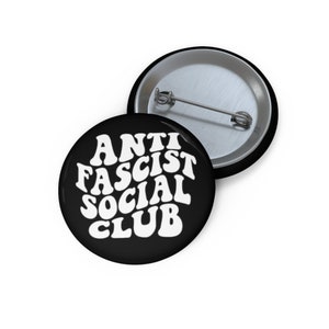 May include: A black button with white text that reads "Anti Fascist Social Club" in a retro font.