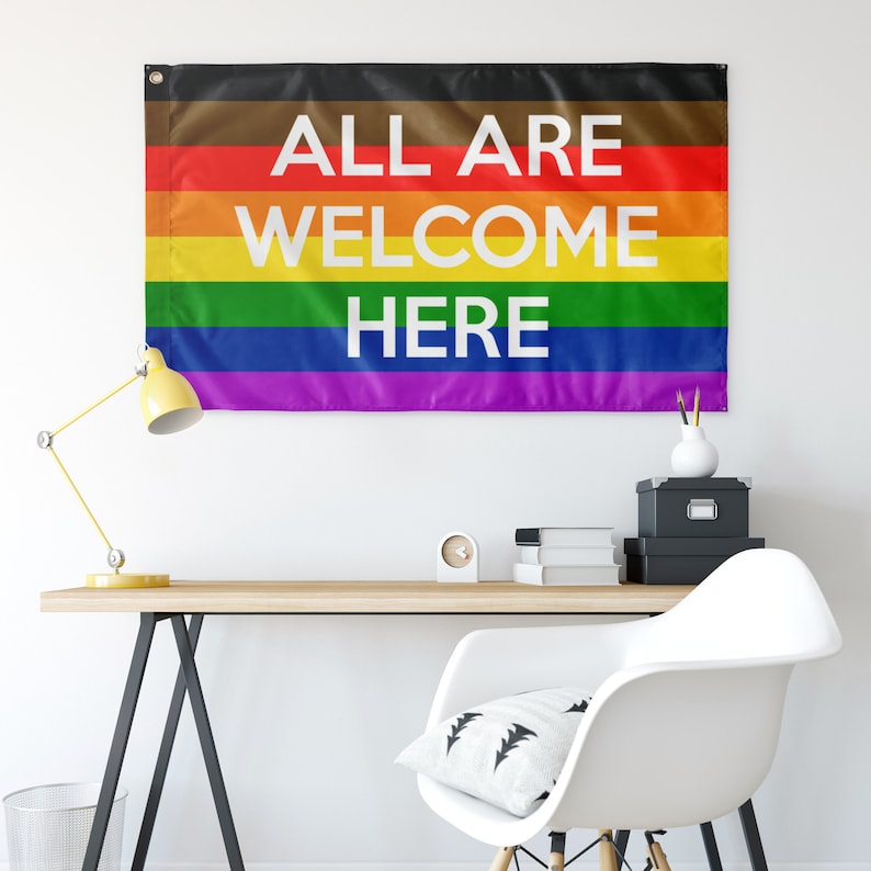 Puede incluir: Una bandera arco&iacute;ris del orgullo con el texto "ALL ARE WELCOME HERE" en letras blancas.