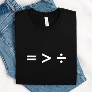 Può includere: Maglietta nera con testo bianco che dice "= > :" su uno sfondo bianco.