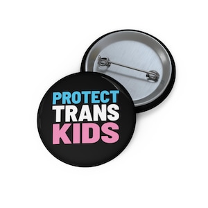 May include: A black button with the text "PROTECT TRANS KIDS" in white, blue, and pink lettering. The button has a silver back with a pin.