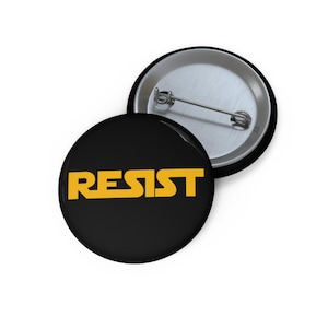 May include: Two round black buttons with the word "RESIST" in yellow block letters. One button is face up, displaying the text, and the other is face down, showing the pin back.