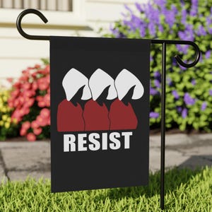 May include: Black garden flag with the word "RESIST" in white block letters. The flag features a graphic of three figures in red robes and white bonnets. The flag is displayed on a black metal stand in a garden.
