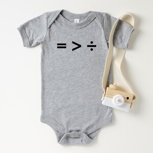 May include: A gray baby onesie with the mathematical symbols: equals, greater than, and division, printed in black. A wooden toy camera with a white front and a cream-colored strap is next to the onesie. The background is white.
