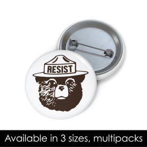 May include: A white button with a brown cartoon image of a bear wearing a ranger hat with the word "RESIST" on it.
