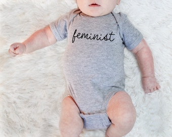 May include: A gray baby onesie with the word "feminist" written in black cursive lettering.