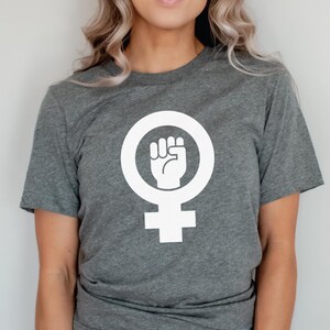 May include: A gray t-shirt with a white feminist symbol featuring a raised fist.