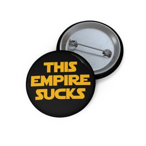 May include: Black button with the text "THIS EMPIRE SUCKS" in yellow, stylized font. The button has a metal pin back. The second button is visible in the background.