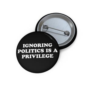 May include: Black button with white text that reads "Ignoring Politics is a Privilege".