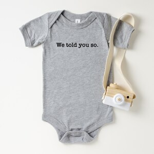 Puede incluir: Un body de bebé gris con el texto "We told you so." Una pequeña cámara de juguete de madera con una correa blanca y beige descansa junto al body. La cámara tiene una parte delantera blanca y un cuerpo beige.