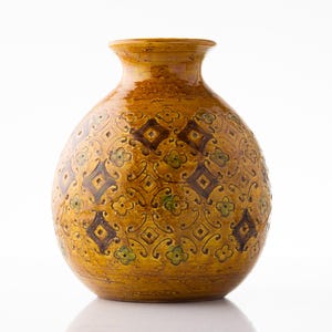 Op de afbeelding: Een decoratieve, handgemaakte keramische vaas met een bolvormige body en een smalle hals. De vaas is voornamelijk mosterdgeel met een herhalend patroon van bruine ruiten en groene bloemaccenten. Voor interieurdecoratie.