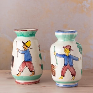 Könnte beinhalten: Zwei handbemalte Keramikvasen. Jede Vase hat eine weiße Basis mit einer farbenfrohen Illustration einer Person mit Hut, blauer Jacke und roter Hose. Die Vasen haben grüne und blaue Ränder und stehen auf einer Holzoberfläche.