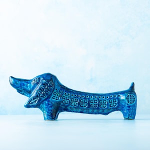 May include: A ceramic dachshund figurine in a vibrant blue glaze. The dog has detailed patterns on its body and head, with a small tail and four legs. The figurine is a decorative item.
