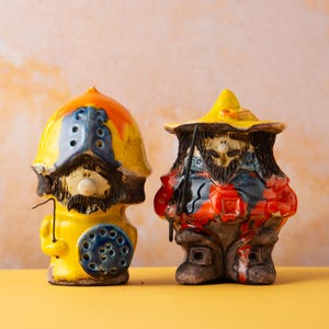Puede incluir: Dos figuras de cerámica caprichosas de hombres barbudos. Una es amarilla, azul y naranja, sosteniendo un alambre. La otra es roja, azul y marrón, con un sombrero y un bastón. Ambas tienen rasgos faciales detallados y están sobre una superficie amarilla.
