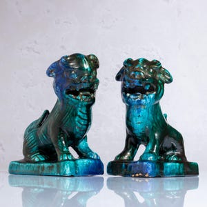 Op de afbeelding: Twee keramische Foo Dog-beelden in zittende positie. De beelden zijn geglazuurd in tinten van teal, blauw en groen, met een glanzende afwerking. Elke hond zit op een rechthoekige basis. De beelden hebben gedetailleerde kenmerken.