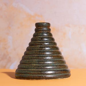 Może przedstawiać: Ręcznie wykonany, ciemnozielony ceramiczny wazon o unikalnym, warstwowym wzorze. Wazon ma szeroką podstawę, która zwęża się ku górze, a każdy poziom jest nieco mniejszy od poprzedniego. Powierzchnia ma teksturowany, nakrapiany wygląd.