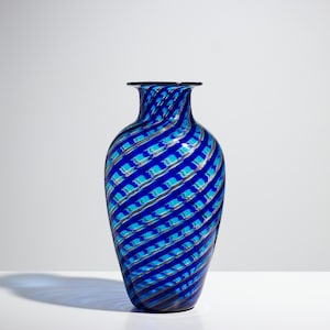 Peut inclure: Un vase en verre présentant une palette de couleurs bleu et turquoise vibrantes avec un motif en spirale diagonal distinct. La surface est ornée de fins rubans argentés métallisés qui s'enroulent autour du corps, créant un effet réfléchissant.
