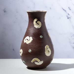 Könnte beinhalten: Eine handgefertigte braune Keramikvase mit breitem Boden und schmalem Hals. Die Vase ist mit cremefarbenen, strukturierten Wirbelmustern und kleinen grünen Akzenten verziert. Die Vase ist etwa 25 cm hoch, perfekt zum Präsentieren von Blumen oder als dekoratives Element.
