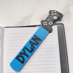 May include: A blue bookmark with the name "DYLAN" printed in black letters. The bookmark is shaped like a video game controller with black and gray details.