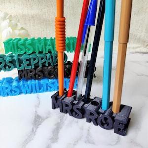 May include: A black plastic pencil holder with the name "MRS ROSE" spelled out in 3D letters. The holder has five slots for pens and pencils. There are five pens and pencils in the holder: a red pencil, a blue pen, a black pen, a blue pen, and an orange pen.