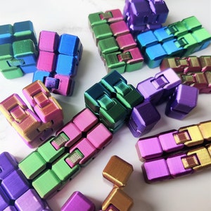 May include: A collection of colourful fidget toys made of metal. The toys are in various shapes and sizes, with some featuring a metallic sheen. The toys are arranged on a white surface.
