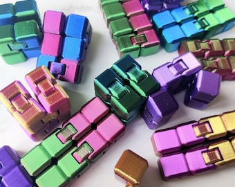 Peut inclure: Une collection de jouets sensoriels colorés en métal. Les jouets sont de formes et de tailles variées, certains présentant un aspect métallique. Les jouets sont disposés sur une surface blanche.