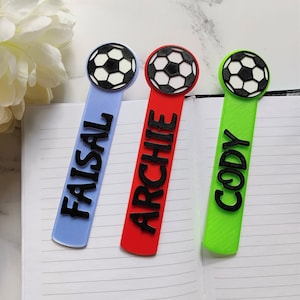 May include: Three personalised bookmarks with football ball toppers. The bookmarks are blue, red, and green with the names "Faisal", "Archie", and "Cody" printed in black.