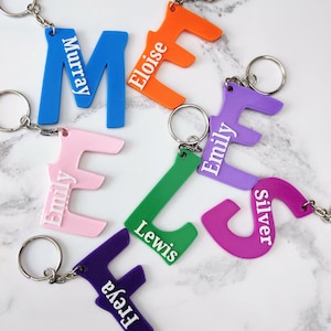 May include: A collection of colorful, personalized keychains featuring the letters 'M', 'E', 'E', 'L', 'S', and 'F' with names printed on each. The keychains are made of plastic and have a silver metal ring.