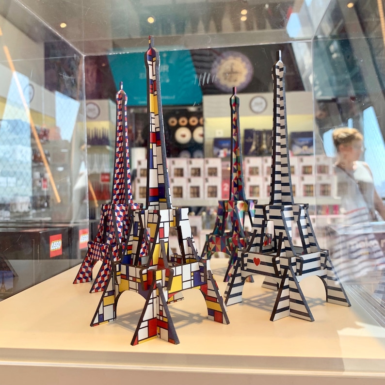 May include: Five miniature Eiffel Towers made of cardboard, each with a different geometric pattern. The towers are painted in various colors, including red, blue, yellow, black, and white. The towers are arranged on a white surface.