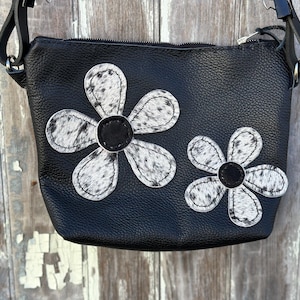 May include: Black crossbody bag with two large, white and black cowhide flower appliqués. The bag has a zipper closure and a black strap. The bag is made of a textured leather material.