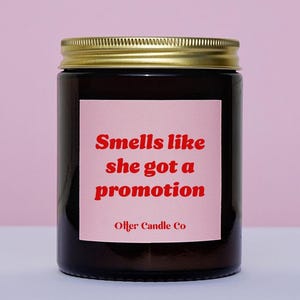 May include: A brown glass candle jar with a gold lid. The label is pink with red text that reads "Smells like she got a promotion" and "Otter Candle Co."