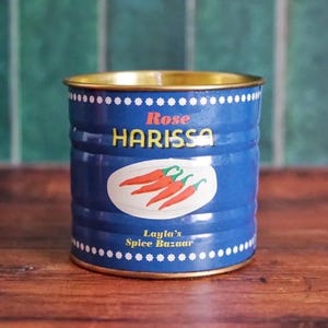 May include: A blue tin with gold edging, labelled "Rose Harissa" and "Layla's Spice Bazaar." The can has a white oval with red chilli pepper illustrations. It sits on a wooden surface against a green wall.