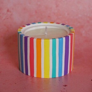 May include: A white candle with a rainbow striped design. The candle is in a cylindrical container with vertical stripes of purple, blue, green, yellow, orange, and red.