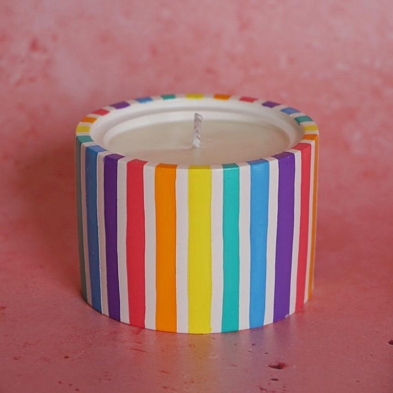 May include: A white candle with a rainbow striped design. The candle is in a cylindrical container with vertical stripes of purple, blue, green, yellow, orange, and red.