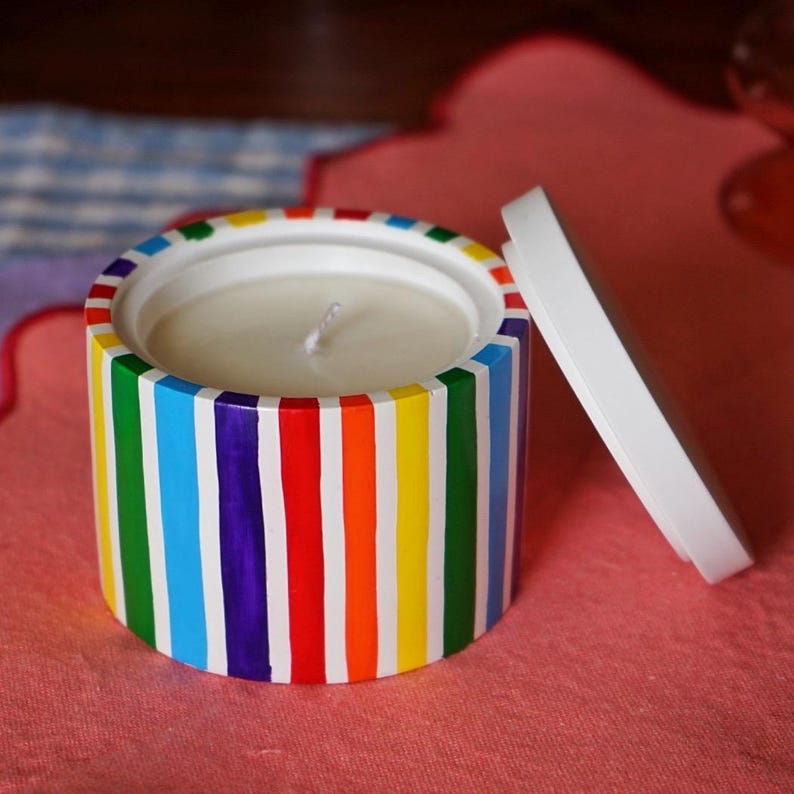 May include: A white candle jar with a rainbow striped design. The lid is off and the candle is white.