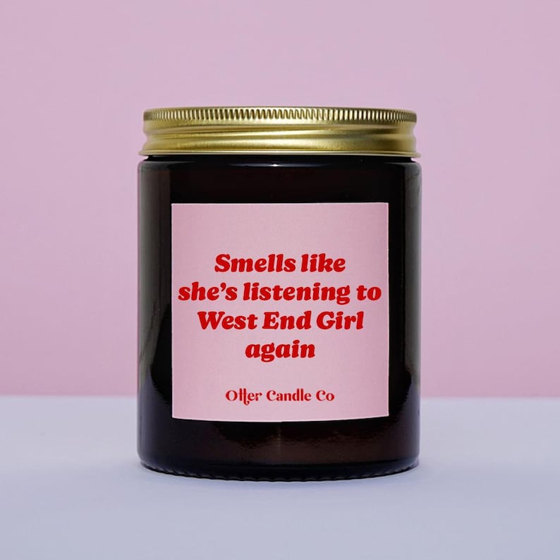 May include: A dark glass jar candle with a gold lid. The label is pink with red text that reads "Smells like she's listening to West End Girl again" and "Olfer Candle Co."